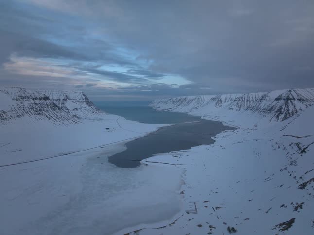 A serene aerial view slowly pulls back to reveal a stunning snow-covered fjord and mountains under a cloudy twilight sky