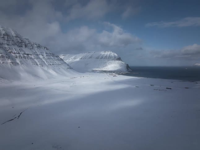 A majestic, static wide shot captures a remote coastal town nestled between vast, snow-covered mountains and the sea