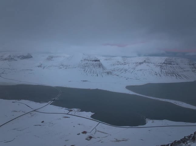 A slow aerial dolly shot reveals a vast, snow-covered mountain range and fjord on a cold, overcast day in Iceland