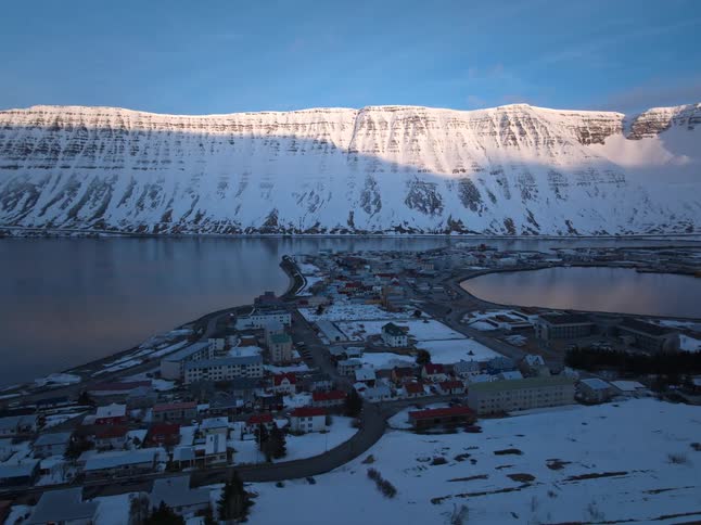 A slow aerial pullback showcases a serene coastal town nestled in a snowy fjord as the morning sun illuminates the towering mountain peaks