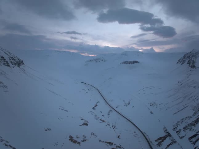 A drone flies forward over a lonely road winding through a spectacular snow-covered mountain range at dusk