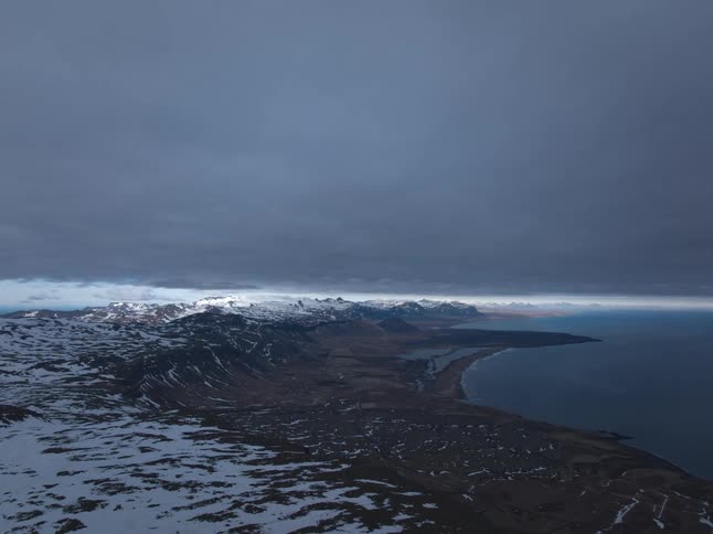 Dramatic storm clouds roll over a snowy Icelandic mountain range next to the sea in a breathtaking time-lapse