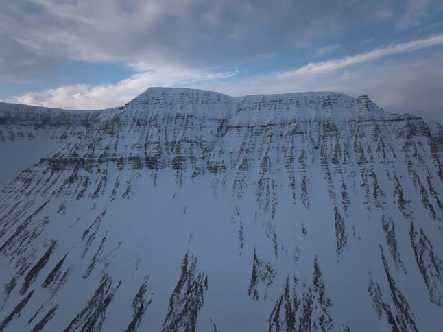An aerial view moves toward and pans across a majestic, snow-covered mountain range in a vast, remote winter landscape