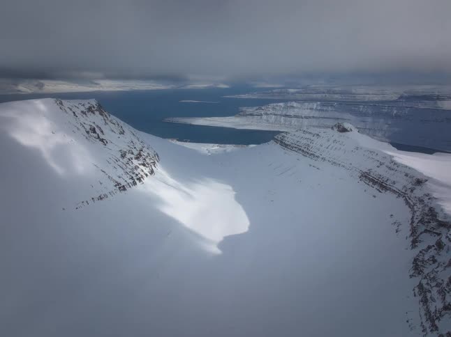 A breathtaking aerial flight over vast snow-covered mountains towards a distant fjord under dramatic clouds
