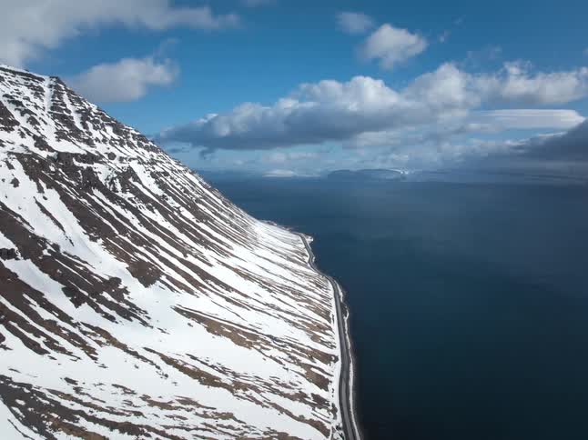 An epic aerial view shows a snow-covered mountain sloping down to meet the dark ocean along a narrow coastal road