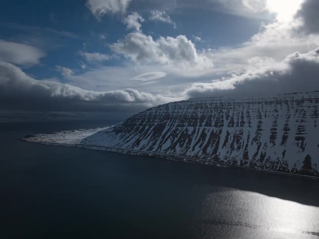 A dramatic aerial time-lapse shows clouds and sunlight moving over a vast, snow-covered mountain range bordering a fjord and a small coastal town