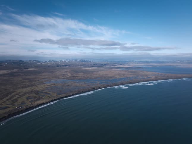 Expansive aerial view of a rugged coastline where the ocean meets a vast marshland with snow-capped mountains in the distance