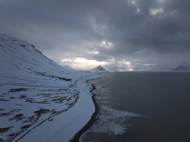 An epic aerial view of a snow-covered Icelandic coastline where dramatic clouds part to reveal stunning sun rays over the ocean