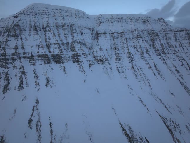 An aerial drone slowly pushes in towards a massive, rugged mountain covered in snow during the cold blue hour