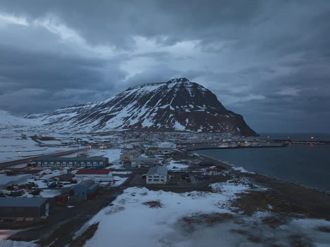 A slow aerial flyover reveals a remote Icelandic coastal town nestled at the base of a snow-covered mountain on a moody winter evening
