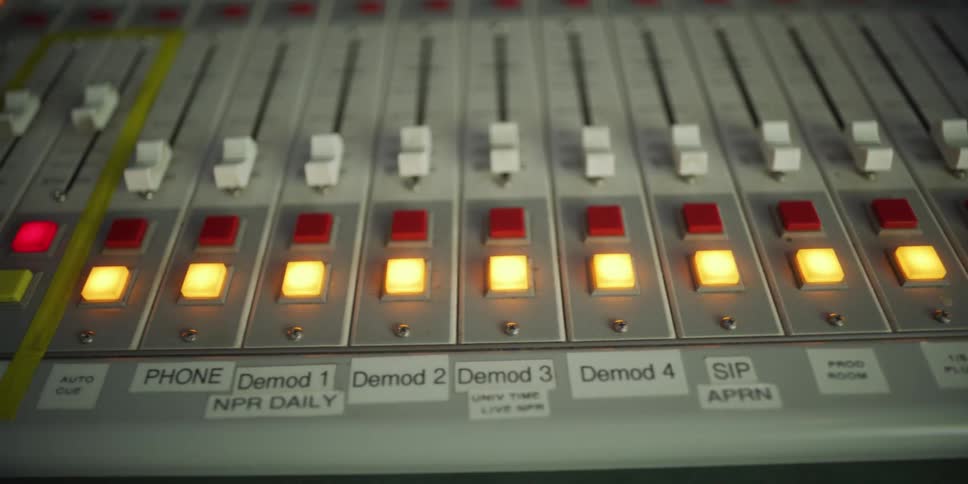 A slow pan shot reveals the illuminated controls of a sound mixing board in a broadcast studio