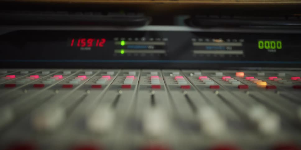 A slow, focused push-in shot of a professional audio mixing console in a recording studio, highlighting the blinking lights, faders, and active VU meters