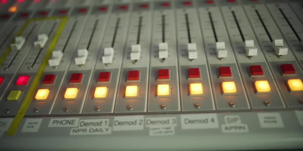 Slow pan across a professional audio mixing console with illuminated buttons in a broadcast studio