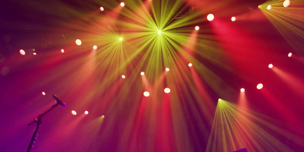 Vibrant yellow and red stage lights illuminate a concert venue with dynamic beams through a hazy atmosphere