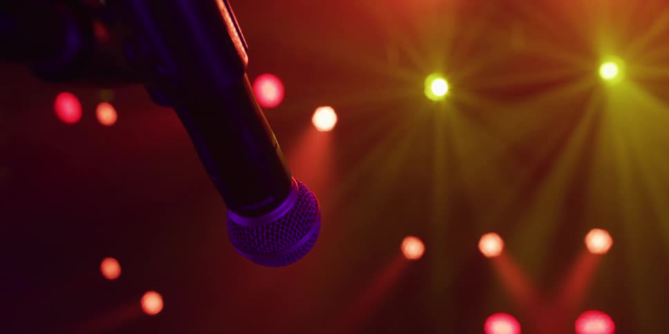 A microphone on a stand awaits the performer under colorful, hazy stage lights at a concert venue