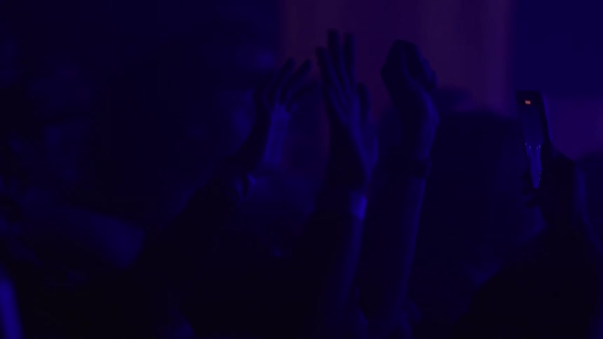 A silhouetted crowd raises their hands and phones to record a performance during a concert with purple lights