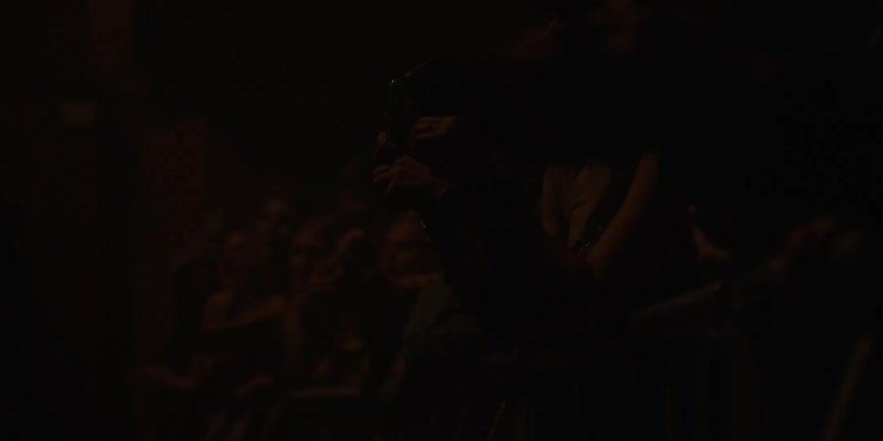 Excited fans in a dark concert venue clap and record the show on their phones in slow motion