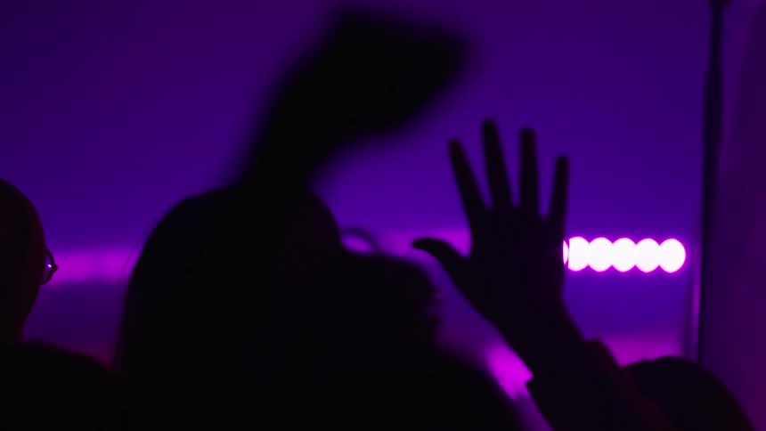 Silhouettes of a crowd worshiping with hands raised at a concert under purple lights in slow motion