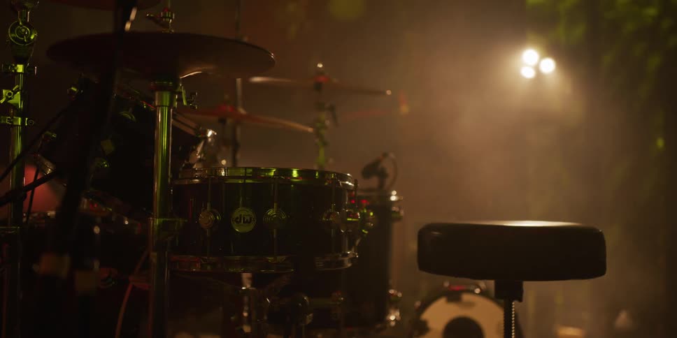 A drum kit sits on a dark, smoky stage as colorful spotlights shift and create a dramatic atmosphere before a show