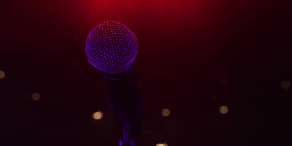 A microphone stands alone on a dark stage, illuminated by moody red and purple lights, waiting for the show to begin