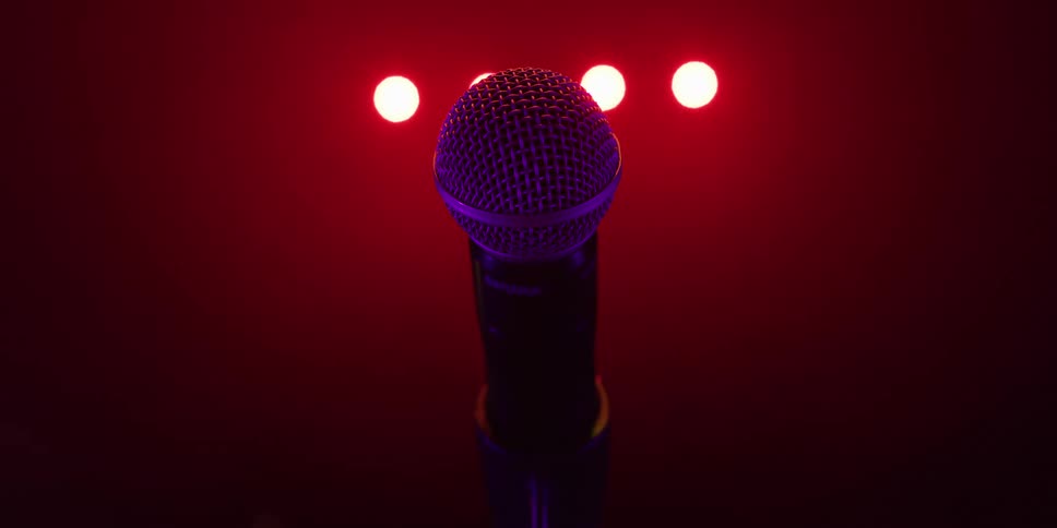 A lone microphone on a stand is illuminated by dramatic red stage lights as the camera slowly zooms in