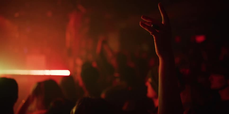 A hand rises in slow motion above a dancing crowd in a dark, hazy club illuminated by flashing green and red lights