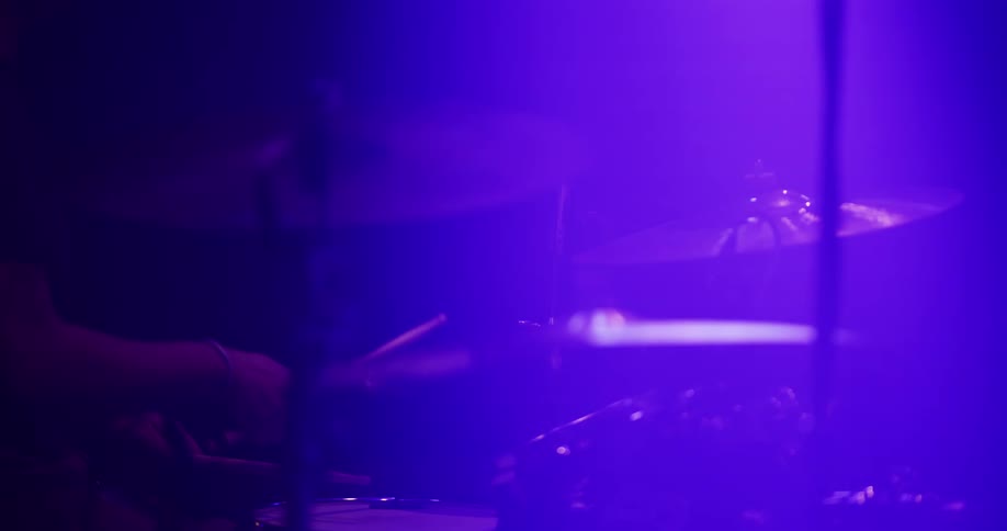 A drummer plays a steady beat on a drum kit, silhouetted against hazy, vibrant purple stage lighting during a live performance