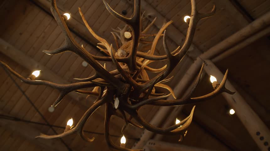 A large, rustic chandelier made from intertwined antlers with lit bulbs hangs from a wooden ceiling in a lodge