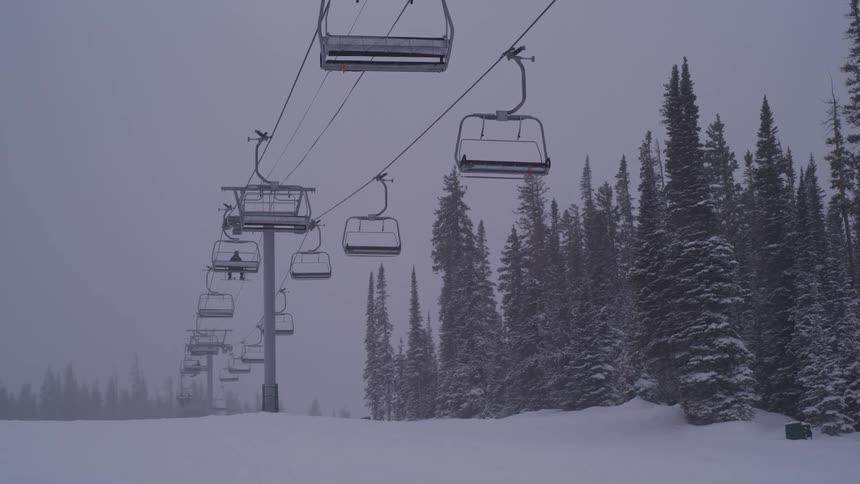 A ski lift with empty chairs moves slowly up a mountain during a quiet snowstorm at a desolate resort