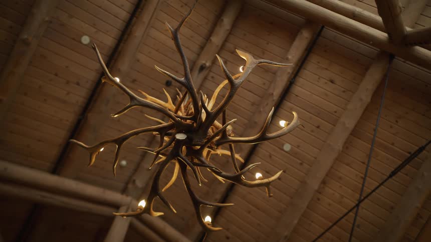 A low-angle shot slowly pulls back from a rustic chandelier made of antlers hanging from the high wooden ceiling of a log cabin