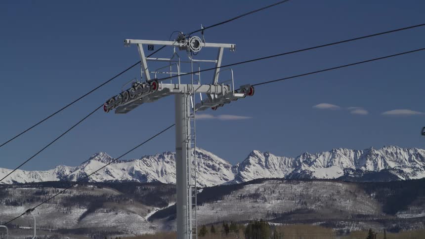 Close-up of a ski lift tower with skiers and snowboarders riding up the mountain against a backdrop of a majestic snowy mountain range