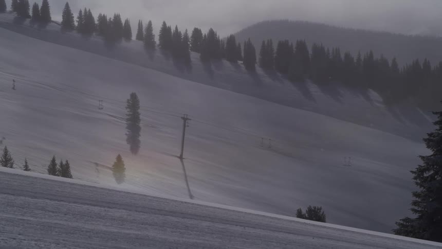 A sun dog appears on the snowy slopes of a quiet, foggy mountain with empty ski lifts during a gentle snowfall