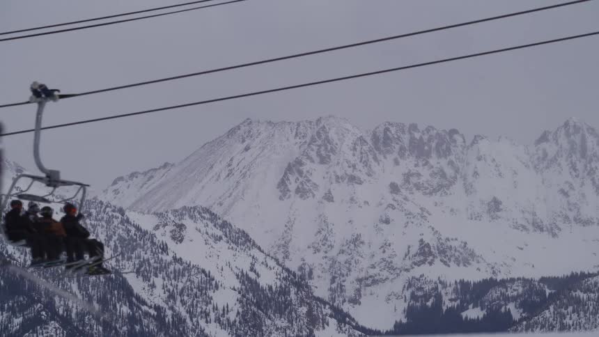 Skiers ascend a snow-covered mountain on a chairlift, with majestic peaks visible in the background