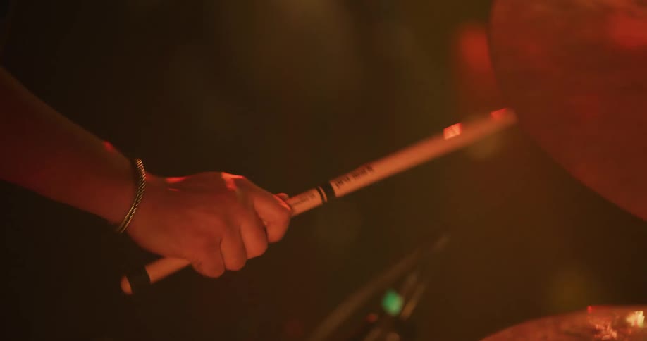 A drummer's hands strike the cymbals in a rhythmic beat in slow motion under colorful, dramatic stage lighting