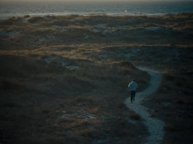 A man jogs along a winding sandy path through coastal dunes towards the ocean at dusk