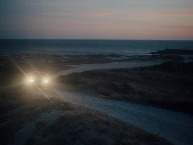A car with its headlights on drives up a winding dirt road through sand dunes along the coast at dusk