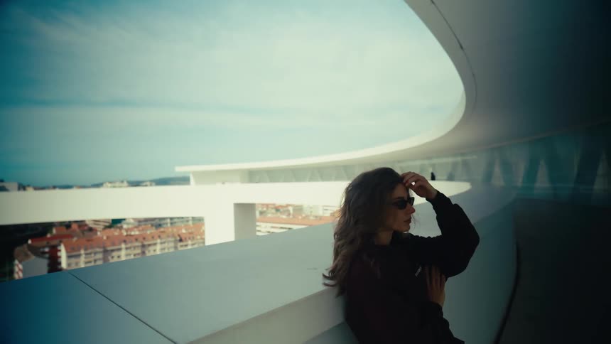 A stylish young woman in sunglasses leans against a modern white architectural structure, thoughtfully gazing at the cityscape below