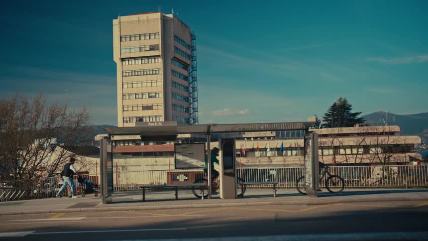 A time-lapse captures the daily city flow as pedestrians, cyclists, and cars blur past a bus stop on a sunny day