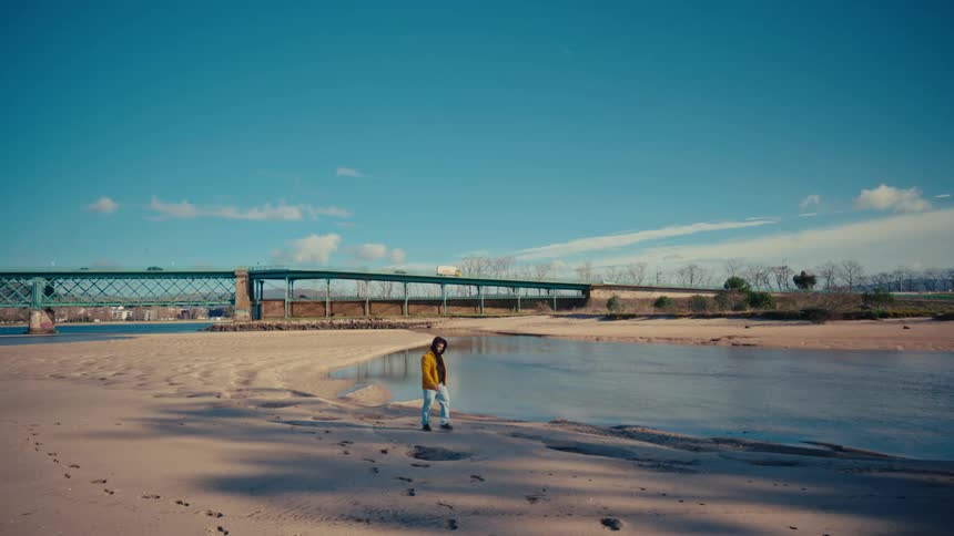 A solitary person in a yellow hooded jacket thoughtfully walks along a sandy riverbank on a beautiful sunny day, with a large bridge in the background