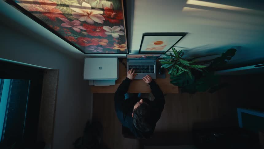 A top-down view shows a man in a hoodie working on his laptop at a modern home office desk, then closing it and walking away