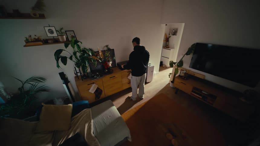 A wide, security camera-style view shows a man putting a vinyl record on a turntable in his dimly lit, modern living room