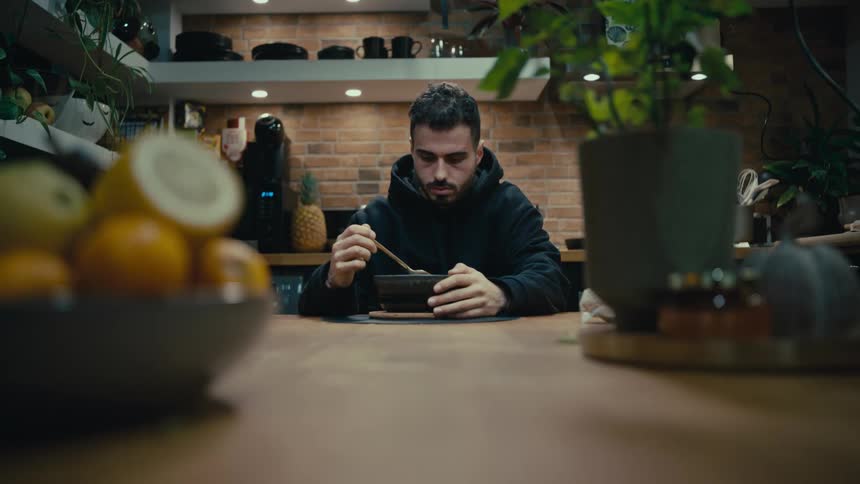 A solitary young man in a black hoodie quietly enjoys a meal from a bowl while seated at a rustic wooden kitchen counter