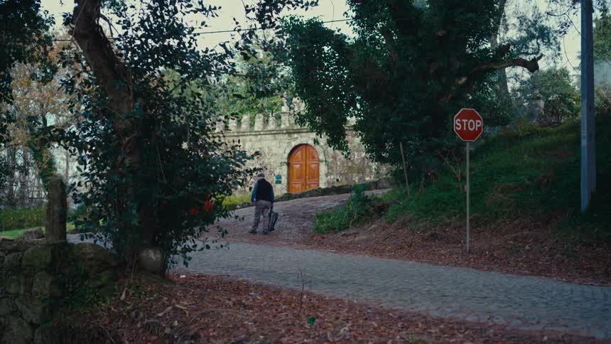 An elderly man pulls a rolling suitcase up a cobblestone path toward a large, historic stone building in the countryside