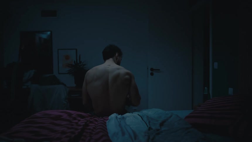 A shirtless and muscular man sits thoughtfully on the edge of his bed in a dark room late at night