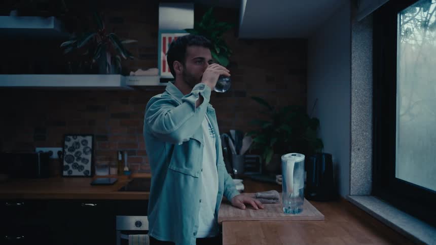A thoughtful young man pours a glass of filtered water in a dimly lit kitchen and drinks it while looking out the window