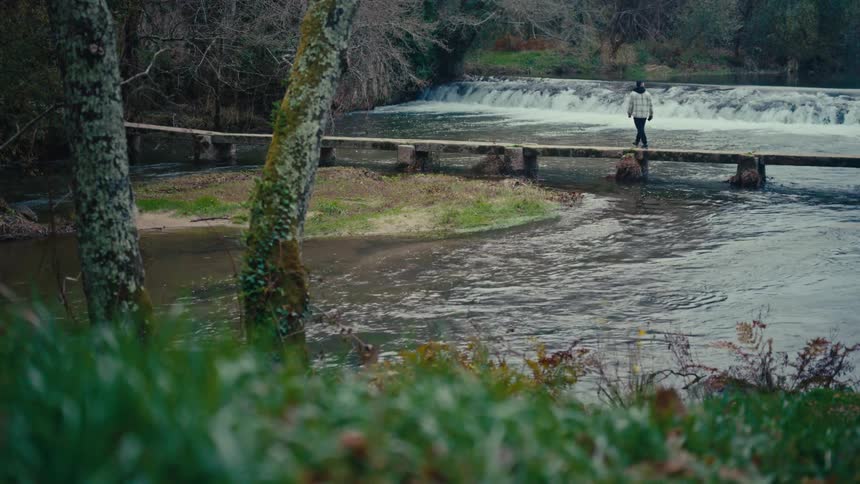 A solitary figure walks back and forth across a rustic stone slab bridge spanning a flowing river near a small waterfall