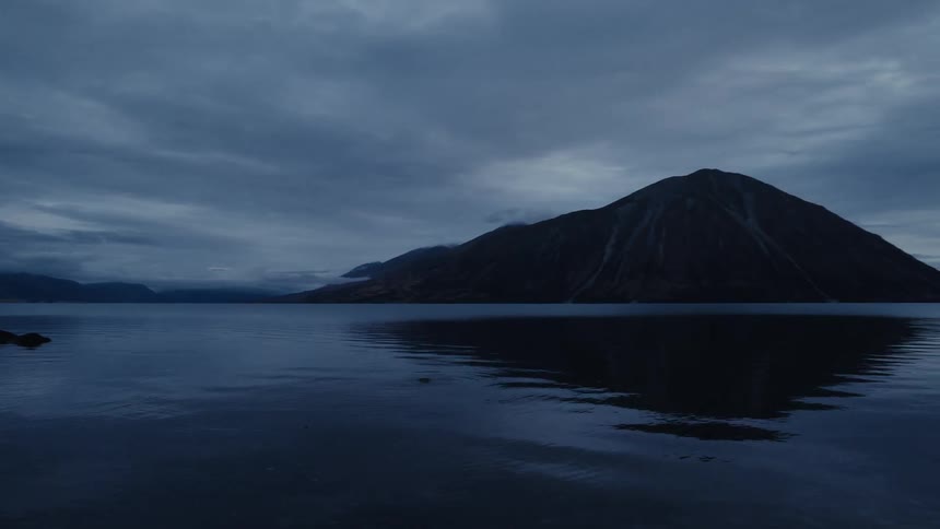 A moody pan across a still mountain lake reveals dramatic reflections of the overcast sky at twilight