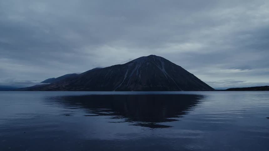 A moody mountain landscape is perfectly reflected in the still, dark water of a lake under a cloudy sky