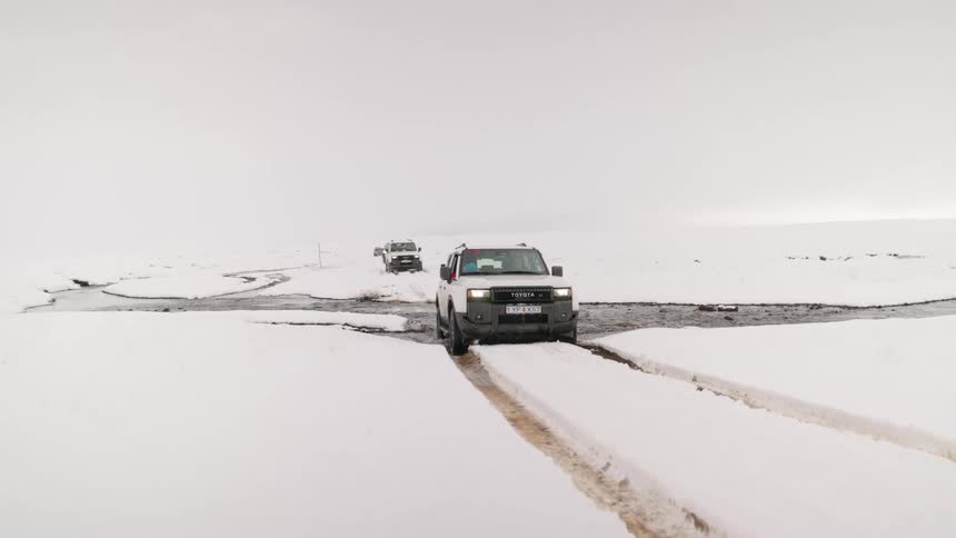A white off-road vehicle leads a convoy through a vast, snowy landscape, driving across a shallow stream