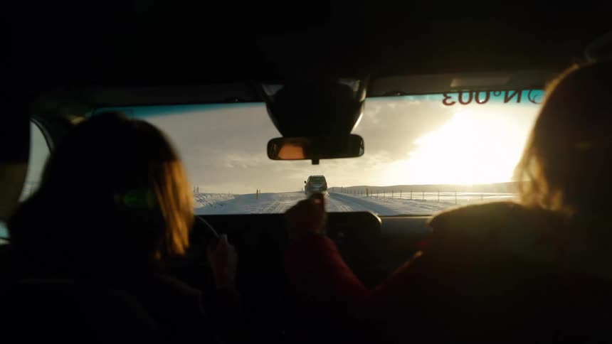 Two women on a winter road trip follow another vehicle down a snowy, remote road into the setting sun